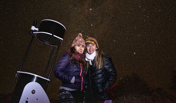 Duas mulheres posando ao lado de um telescópio e com o céu estrelado ao fundo