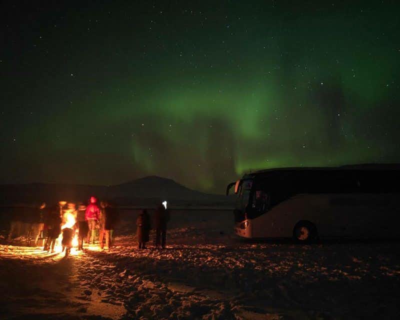 Viajantes ao redor de uma fogueira com a aurora boreal ao fundo